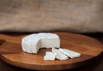 Brie or camembert cheese cut out wedge and in slices on a wooden chopping cutting cheese board on dark brown wooden table with a light brown burlap background