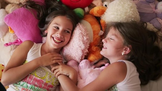 Two little friends chat and laugh, lying in the bed covered of lots of soft toys. Joyful childhood. Friends forever. Moments of happiness. Cute smile. Happy memories. Inside shooting in the nursery.