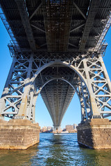 A closeup look up under the Williamsburg Bridge near one of the supporting towers, with Brooklyn in the distance