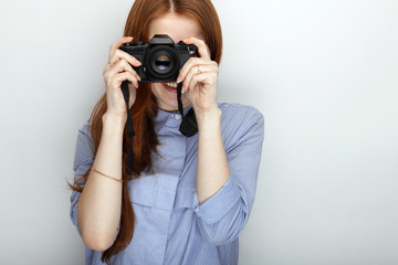 Portrait of cute redhead photographer woman wearing blue striped shirt smiling with happiness and joy while posing with camera against white studio background.