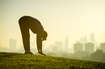 Silhouette of a man stretching on the grassy top of Primrose Hill in front of a misty golden sunrise view of the London city skyline