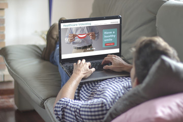 Young man lying on the sofa while using laptop with healthcare videoblog on the screen. View from behind. All screen graphics are made up.