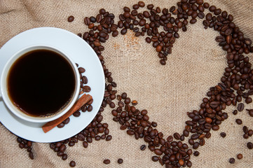 Coffee beans in form of heart and white cup with cinnamon sticks on sackcloth