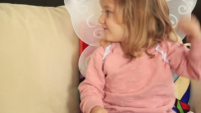 Beautiful little kid with wings, sit and smile on sofa at home interior