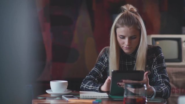 Modern girl in a cafe drinking tea, uses the tablet and writes in notebook. Beautiful girl in the coffee shop working or studying. Remote work. Stylish woman with a tab and notebook in a coffee shop