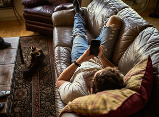 bearded guy relaxing on couch at home using smart phone - obrazy, fototapety, plakaty