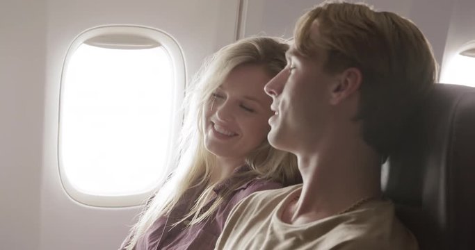 Attractive young couple enjoy each other's company as they fly in main cabin of commercial jet airliner. Close up profile shot, recorded hand-held at 60fps
