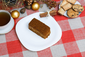 Fototapeta na wymiar Cake on a white plate on a checkered tablecloth with tea, biscuits, cinnamon on a background of a New Year's Christmas tree. New concept. For holiday. View from above.