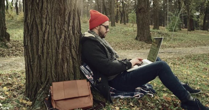 Young man typing on laptop, sitting on ground under tree in park
