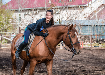 Fototapeta premium Young beautiful brunette girl rides a horse on a warm and sunny autumn day. Portrait of a pretty young woman on the horse, wearing tall boots and gloves.