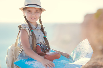 Cute little girl 6 years old,long hair, braided in two braids,wearing a summer Romper sky blue color,sitting on the old suitcase among country roads with an old camera and a map in hand