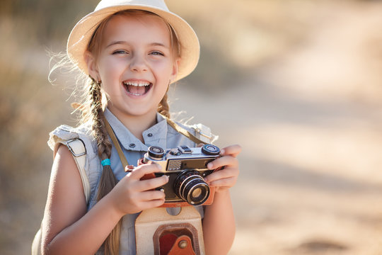 Girl 6 years old with an old brown suitcase and an old camera in hand,brunette with two braids,wearing a straw hat  in a blue flight suit,nature photographs while traveling,standing on a country road