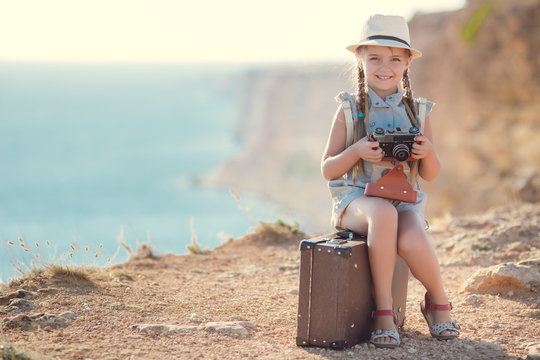 A girl of 6 years with an old old camera,a brunette with two braids,a straw hat, in a blue summer suit,pictures of nature while traveling,sitting on a suitcase on country road