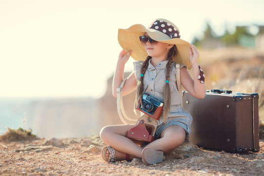 A little girl with pigtails,wearing a straw hat with a large brim,with the old camera around his neck,sitting on top of a cliff,taking pictures of the beautiful nature,the mountains and the sea