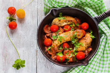 Roasted chicken fillet, cooked with mushrooms, garlic, paprika and olive oil. Cast-iron skillet and fresh cherry tomatoes on wooden table, top view.