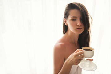 Cute young woman relaxing at cozy atmosphere next to window. Caucasian woman wearing white towel with cup of hot chocolate or coffee in her hands, look at one side. Soft light and natural beauty.