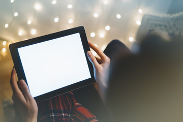 Hipster girl using tablet technology in home atmosphere, person holding computer on background glow bokeh Christmas illumination, female hands texting on relax xmas decoration, mockup templates, blur