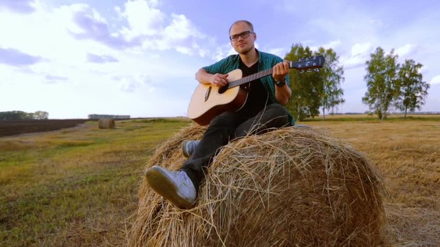 Thoughtful handsome guy playing guitar, sitting on a haystack. The concept of loneliness, happiness and a gay cowboy. Russian field and nature