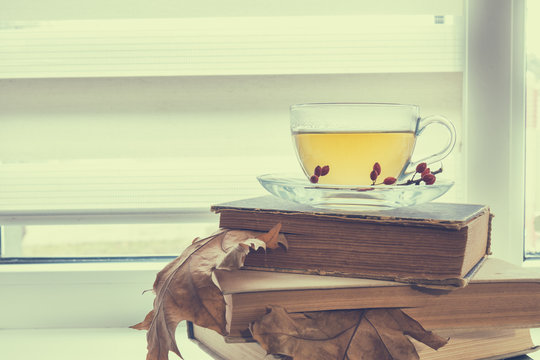 Cup of hot tea, old books, autumn leaves and berries on the window. Autumn still life. Coloring and processing photo.