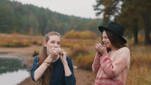 One girl plays harmonica and another girl listens music and drinks hot tea in the autumn forest near the river