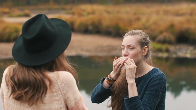 One girl plays harmonica and another girl listens music and drinks hot tea in the autumn forest near the river