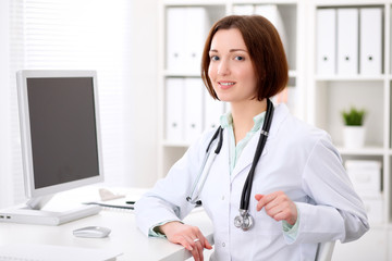 Young brunette female doctor sitting at a desk and working on the computer at the hospital office.  Health care, insurance and help concept. Physician ready to examine patient