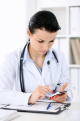 Young brunette female doctor sitting  with clipboard near window in  hospital and filling up medical history form.