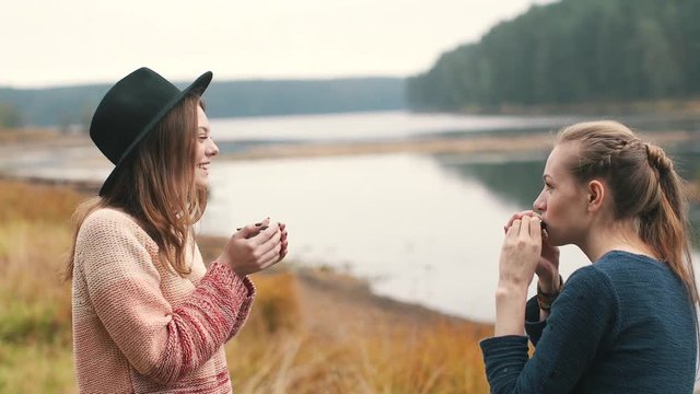 One girl plays harmonica and another girl listens music and drinks hot tea in the autumn forest near the river