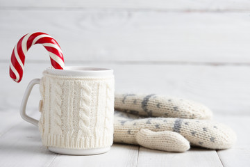 Knitted winter cup of hot drink with a candy cane and mittens on white wooden background