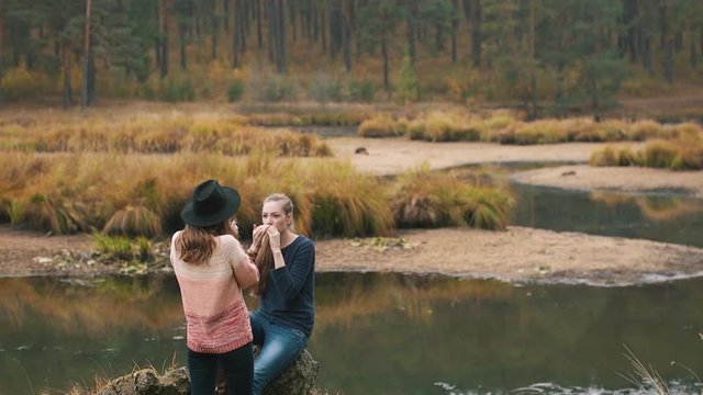 One girl plays harmonica and another girl listens music and drinks hot tea in the autumn forest near the river