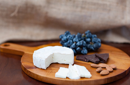 Brie camembert cheese wedge slices almonds, dark chocolate pieces dark blue purple black concord grapes on wooden chopping cheese board dark brown wooden table brown burlap background copy space