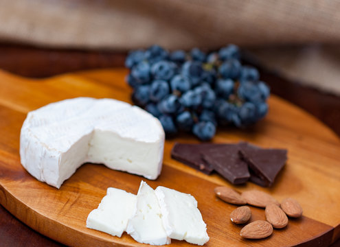 Brie camembert cheese wedge slices almonds, dark chocolate pieces dark blue purple black concord grapes on wooden chopping cheese board dark brown wooden table brown burlap background copy space