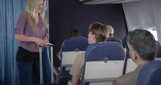 Young man helps young woman stow a heavy carry on bag in main cabin of commercial airliner.  Then they sit down together.  Medium shot from rear of plane.