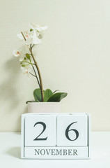 Closeup white wooden calendar with black 26 november word with white orchid flower on white wood desk and cream color wallpaper in room textured background , selective focus at the calendar