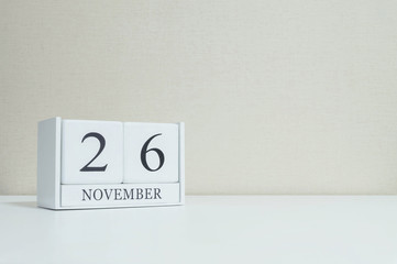 Closeup white wooden calendar with black 26 november word on blurred white wood desk and cream color wallpaper in room textured background with copy space , selective focus at the calendar