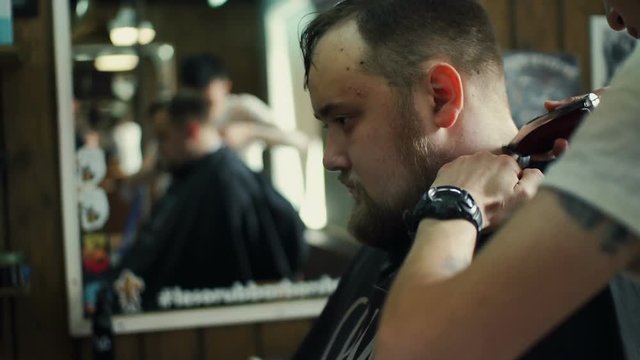 Perfect trim. Rear view close-up of young bearded man getting haircut by hairdresser with electric razor while sitting in chair at barbershop