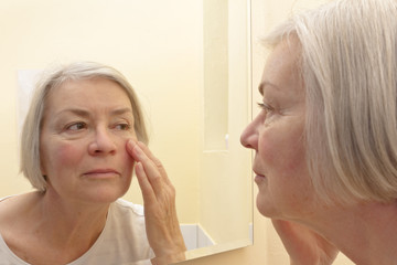 Senior woman with grey hair having a close look at the wrinkles of her facial skin in a mirror, thinking about esthetic surgery