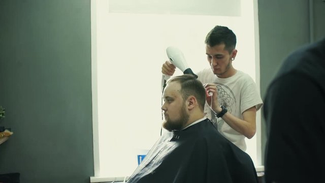Portrait of young bald bearded man getting shaved with straight edge razor by hairdresser at barbershop