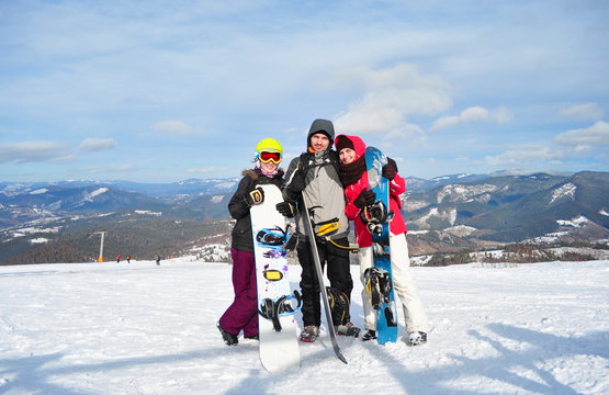 Two girls and a boy standing, smiling and holding snowboards on the background of the Carpathian mountains on a sunny winter day.