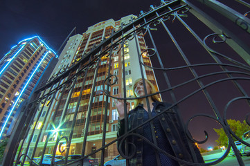 girl in the yard at night. young blond woman standing alone in wrought fence.