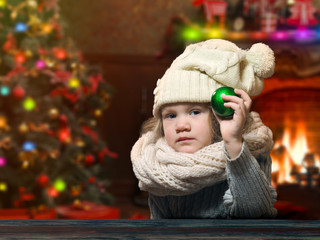 Serious little girl shows a green Christmas ball. House with Christmas decor. Christmas tree, fireplace. Child in hat and scarf