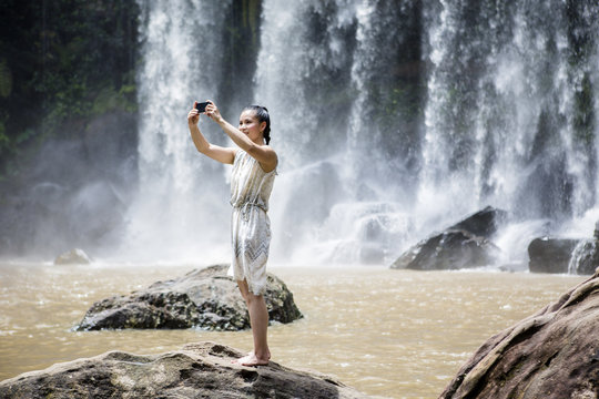 Cambodia, National park Phnom Kulen, young woman taking selfie with smartphone in front of waterfalls