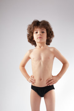 Cute curly-haired boy in shorts without clothes shows her body. Gray background.