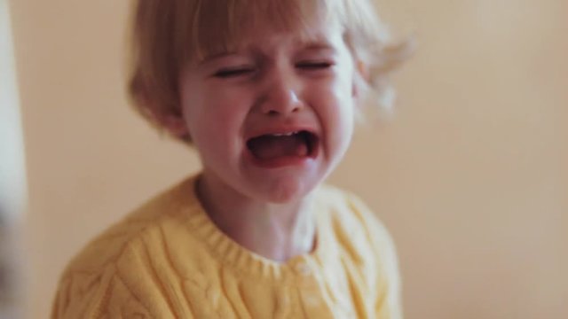 A little cute blonde baby-girl in a yellow warm sweater in the home interior surroundings crying loudly. Angry little girl with sad expression, screaming and crying. Close up view.