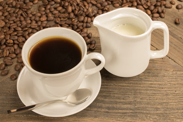 Cup of black aromatic coffee and jug of milk.
In corner coffee beans spread over wooden table.