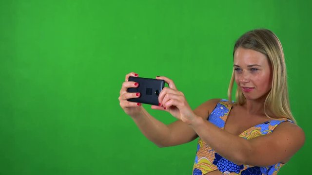 young pretty blond woman takes photos wirh smartphone - green screen - studio 