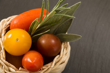Colorful cherry tomatoes in wicker basket with herbs, on black background.