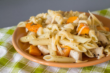 Penne pasta with chicken, pumpkin, basil and cheese in orange plate on checkered tablecloth
