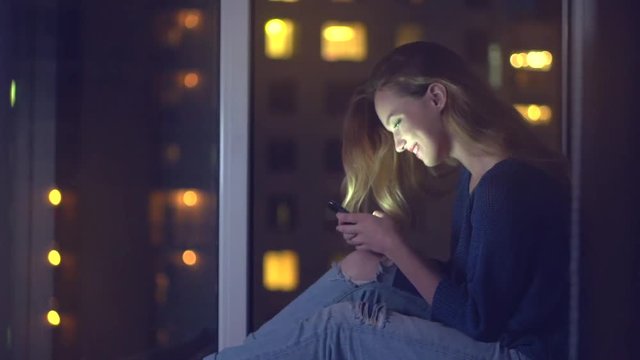 Beautiful girl sitting near the window at home at night and using smartphone. Full HD 1080p video footage