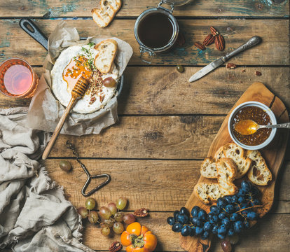 Cheese, fruit and wine set. Camembert in small pan, honey, fig jam, persimmon, grapes, pecan nuts, grilled baguette slices and glass of rose wine over rustic wooden background, top view, copy space